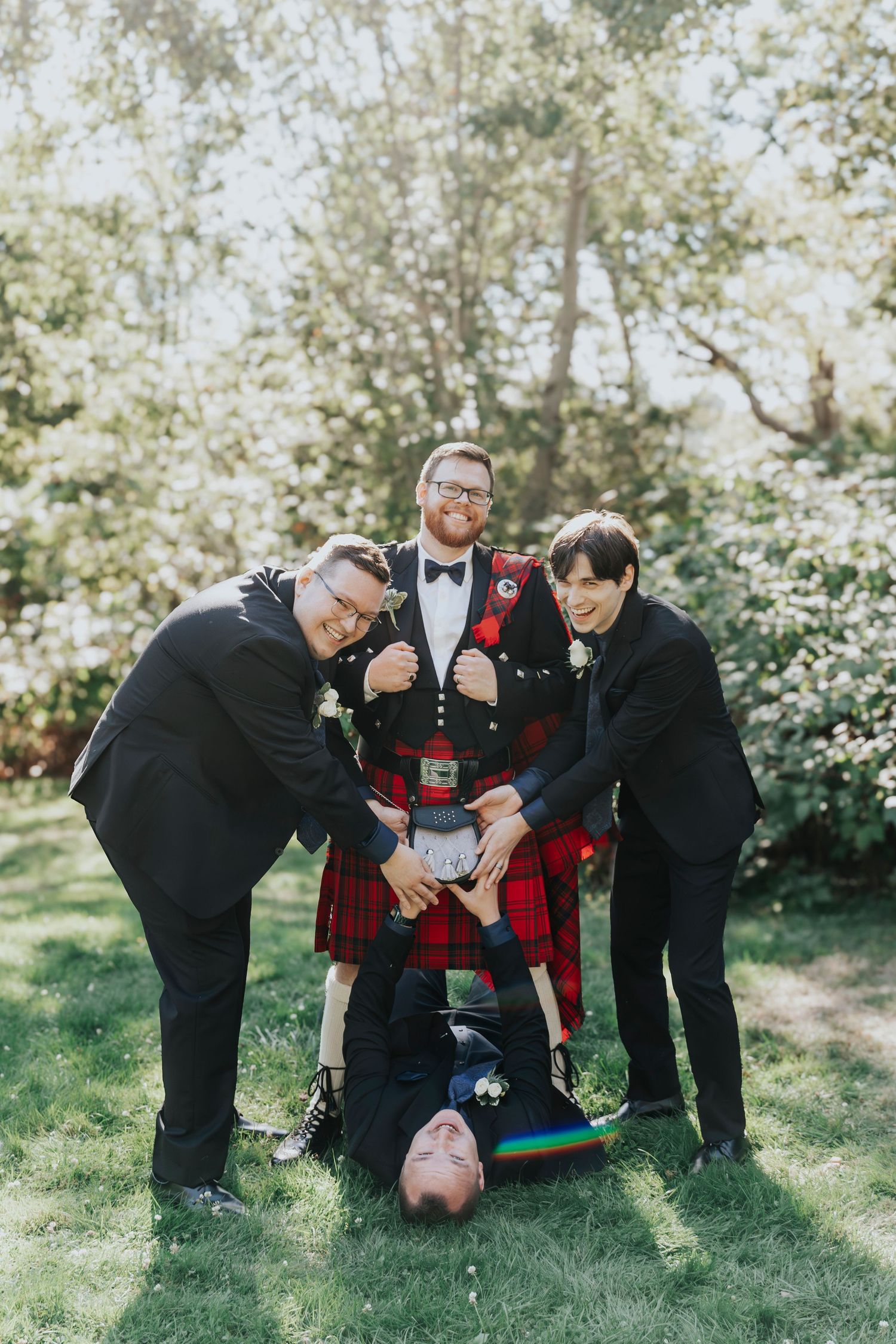 Groom in red tartan kilt with groomsmen in black suits posing playfully outdoors at a wedding.