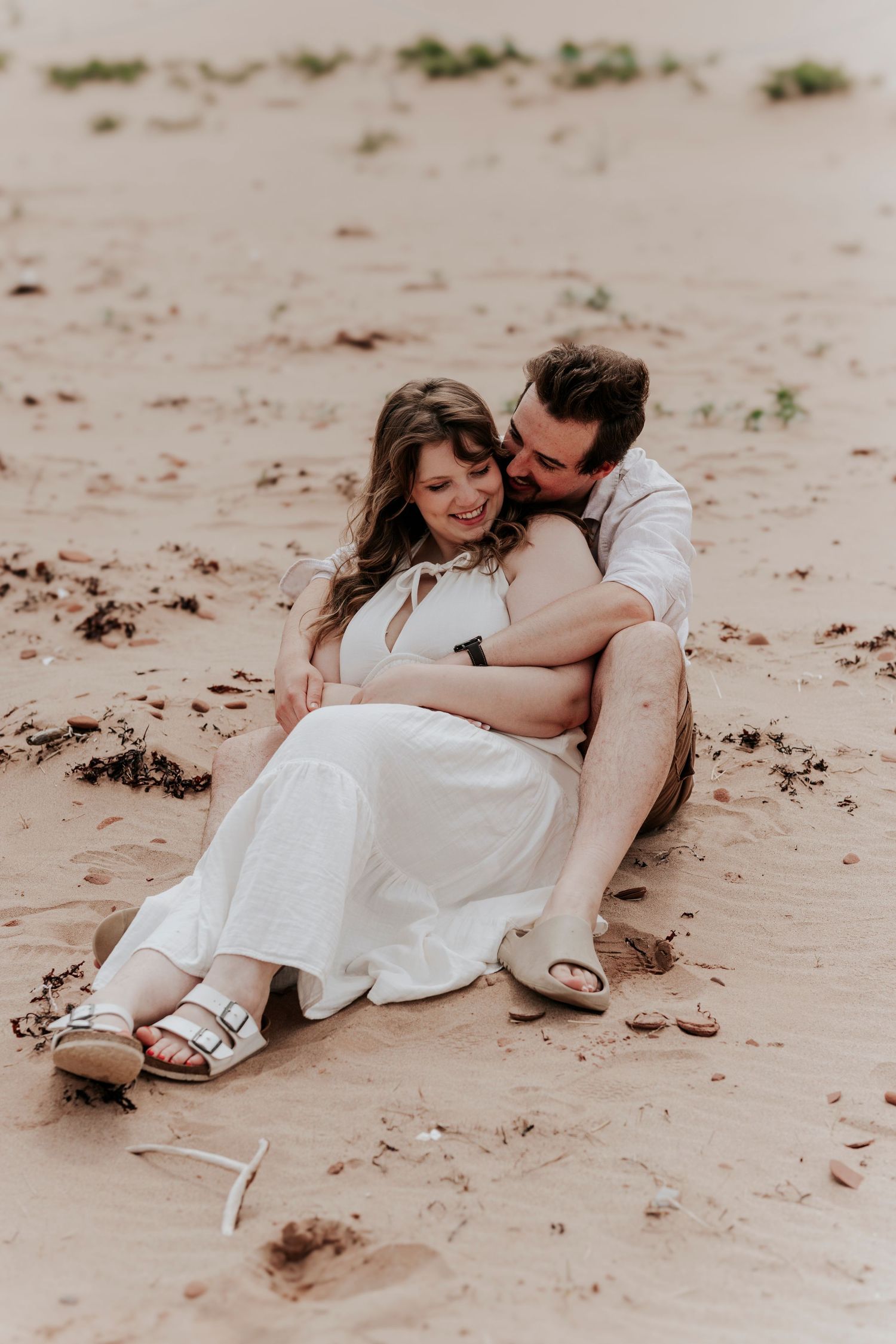 Couple sitting together on sandy beach, man embracing woman in white dress from behind, sharing a tender moment.