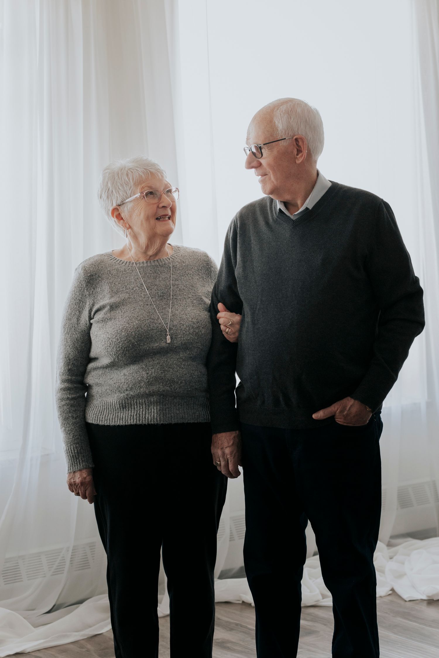Happy elderly couple standing together, looking at each other lovingly near white curtains.