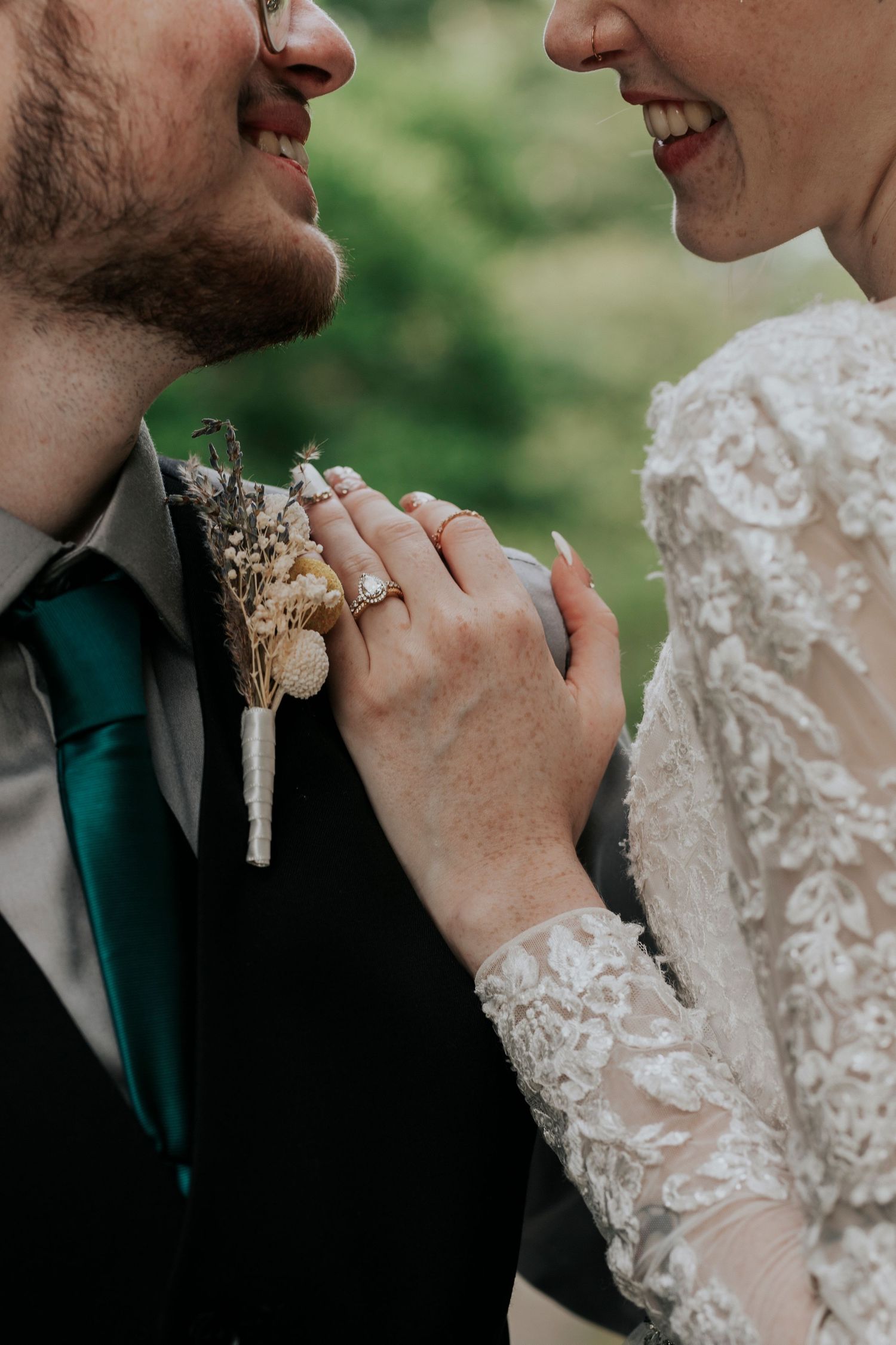 Close-up wedding photo showing clasped hands with engagement ring and dried floral boutonniere against lace dress sleeve.