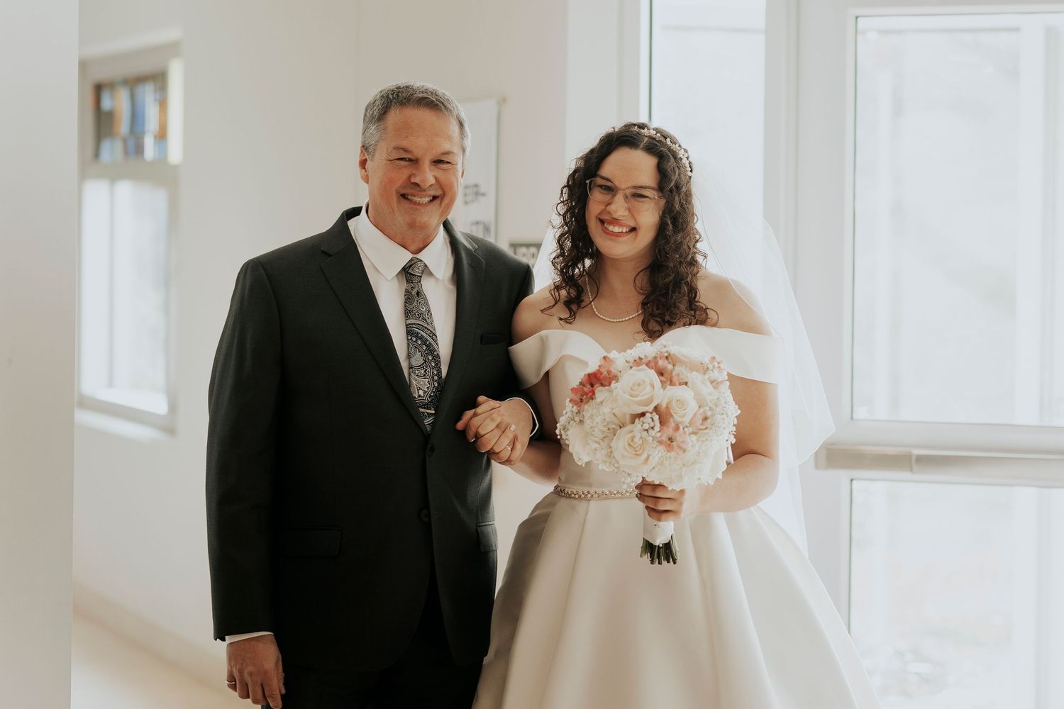 A wedding photo showing two people in formal attire standing in a bright white room with bouquet.