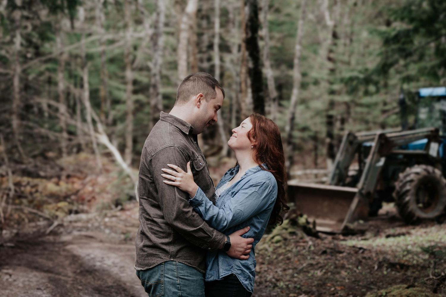 Couple embracing tenderly on a forest path surrounded by tall pine trees and rustic machinery.