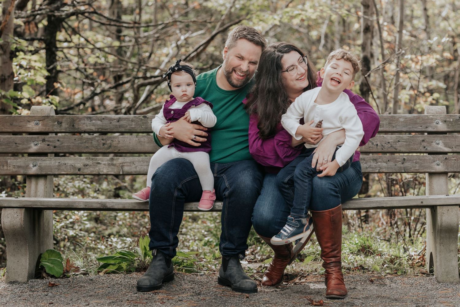 Happy family of four sits together on a wooden bench in an autumn forest, laughing and smiling together.