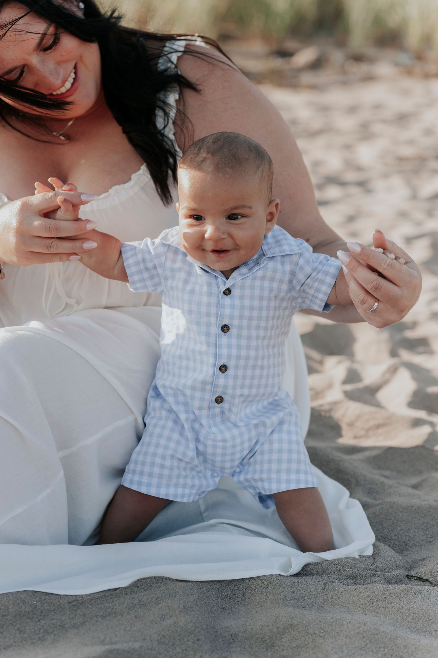 Smiling baby in blue gingham romper sits on sandy beach with mother during family photo session.
