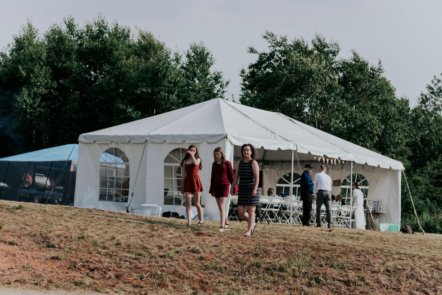 A white event tent set up on a hillside with guests gathering outside during a daytime celebration.