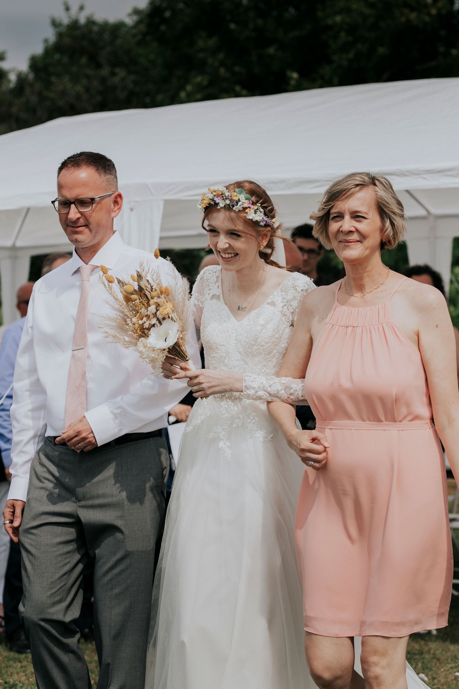An outdoor wedding celebration with a white tent backdrop and guests dressed in formal attire.