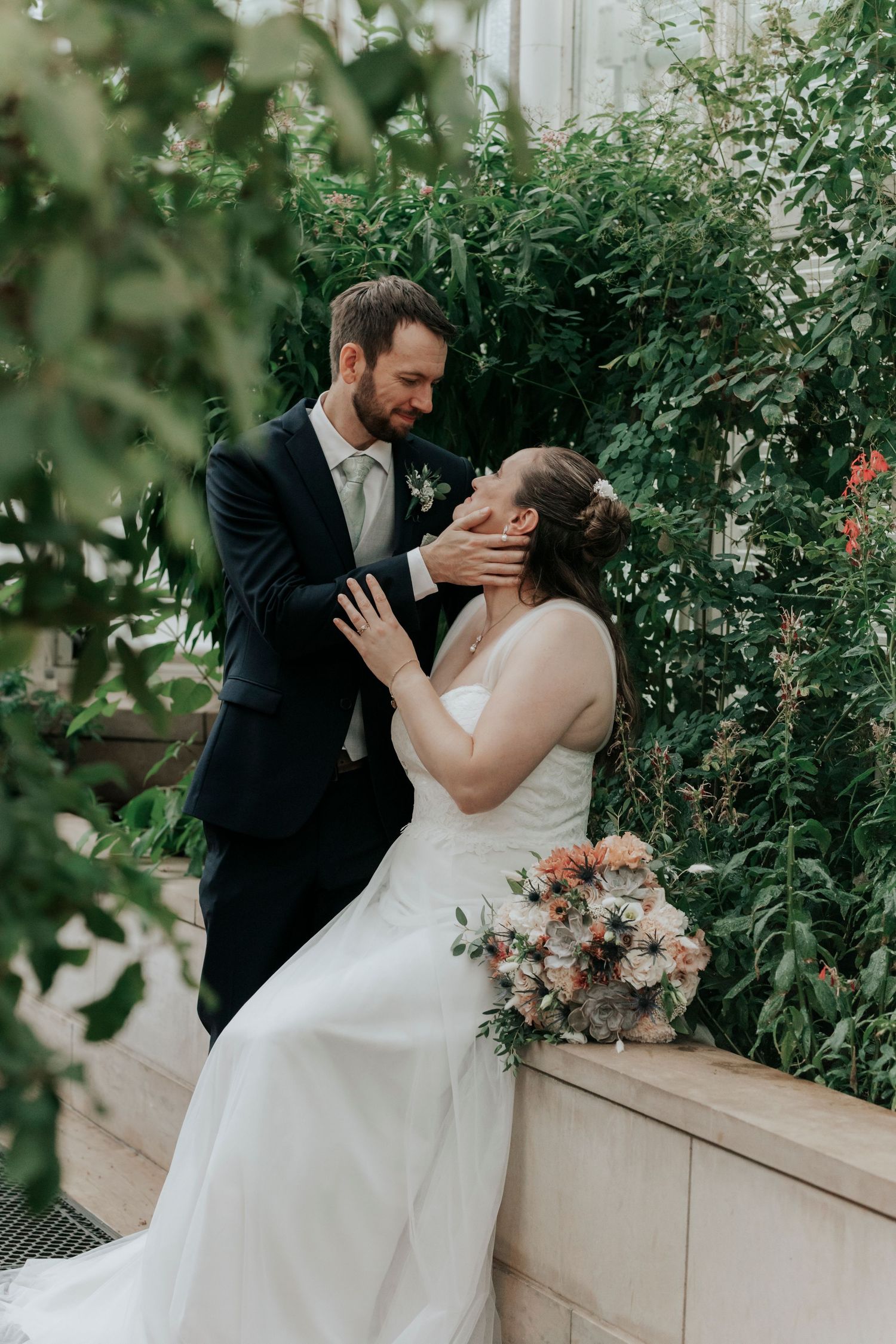 Groom tenderly cups bride's face as she sits holding a bouquet in a lush greenhouse wedding portrait.
