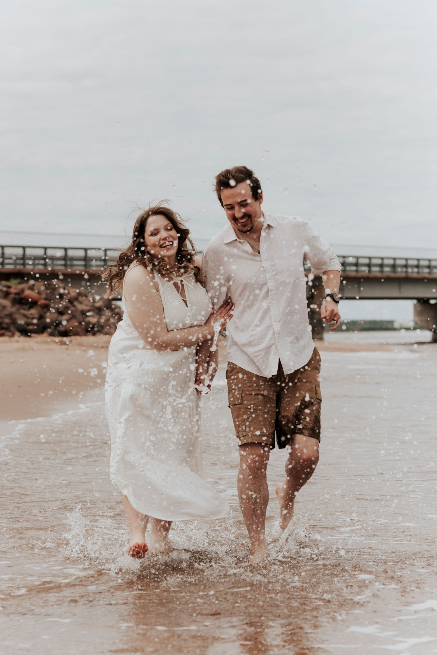 Joyful couple runs through ocean waves on the beach, splashing water, during an engagement photo session.