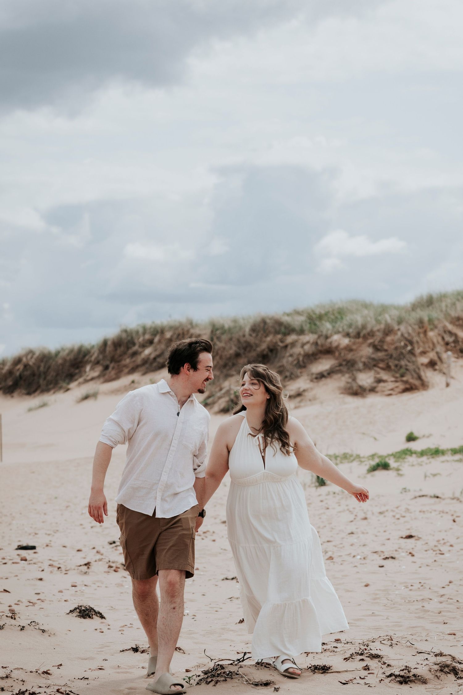 Smiling couple walking hand in hand on sandy beach with sand dunes and dramatic cloudy sky.