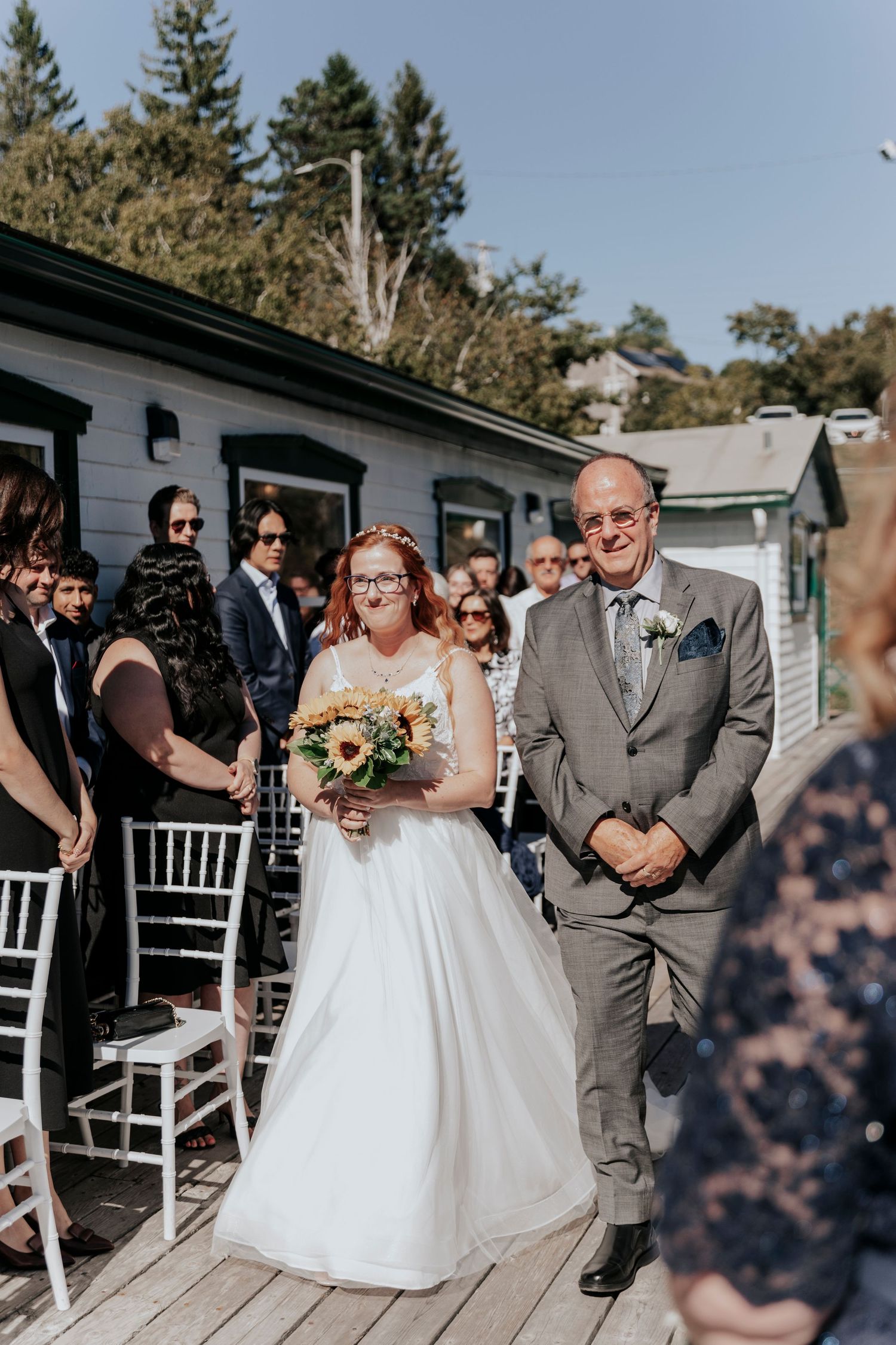 Bride with red hair walks down outdoor aisle holding sunflower bouquet, escorted by father in gray suit.