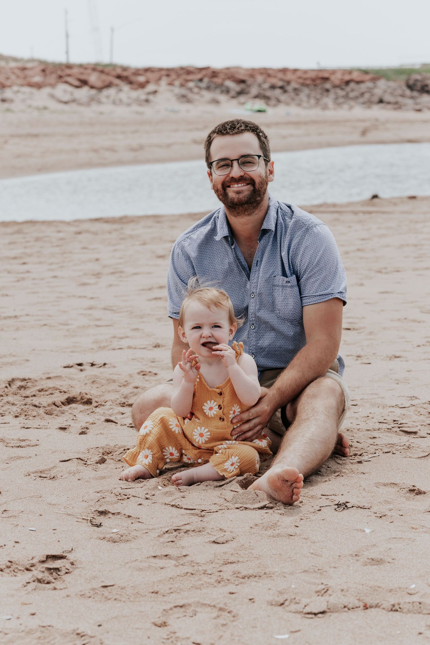 Father and baby daughter sitting together on sandy beach smiling.