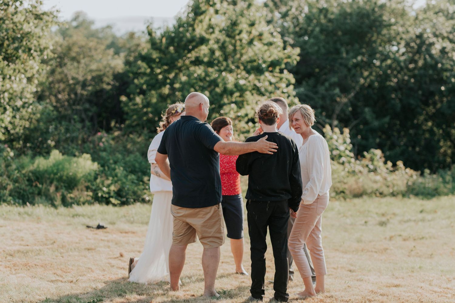 A group of people hugging in a circle outdoors on a sunny day with green trees in the background.
