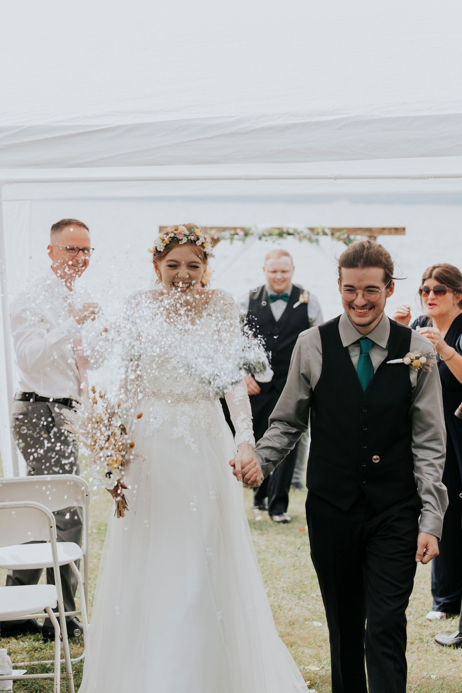Wedding couple walks down aisle while guests throw confetti during outdoor ceremony celebration.