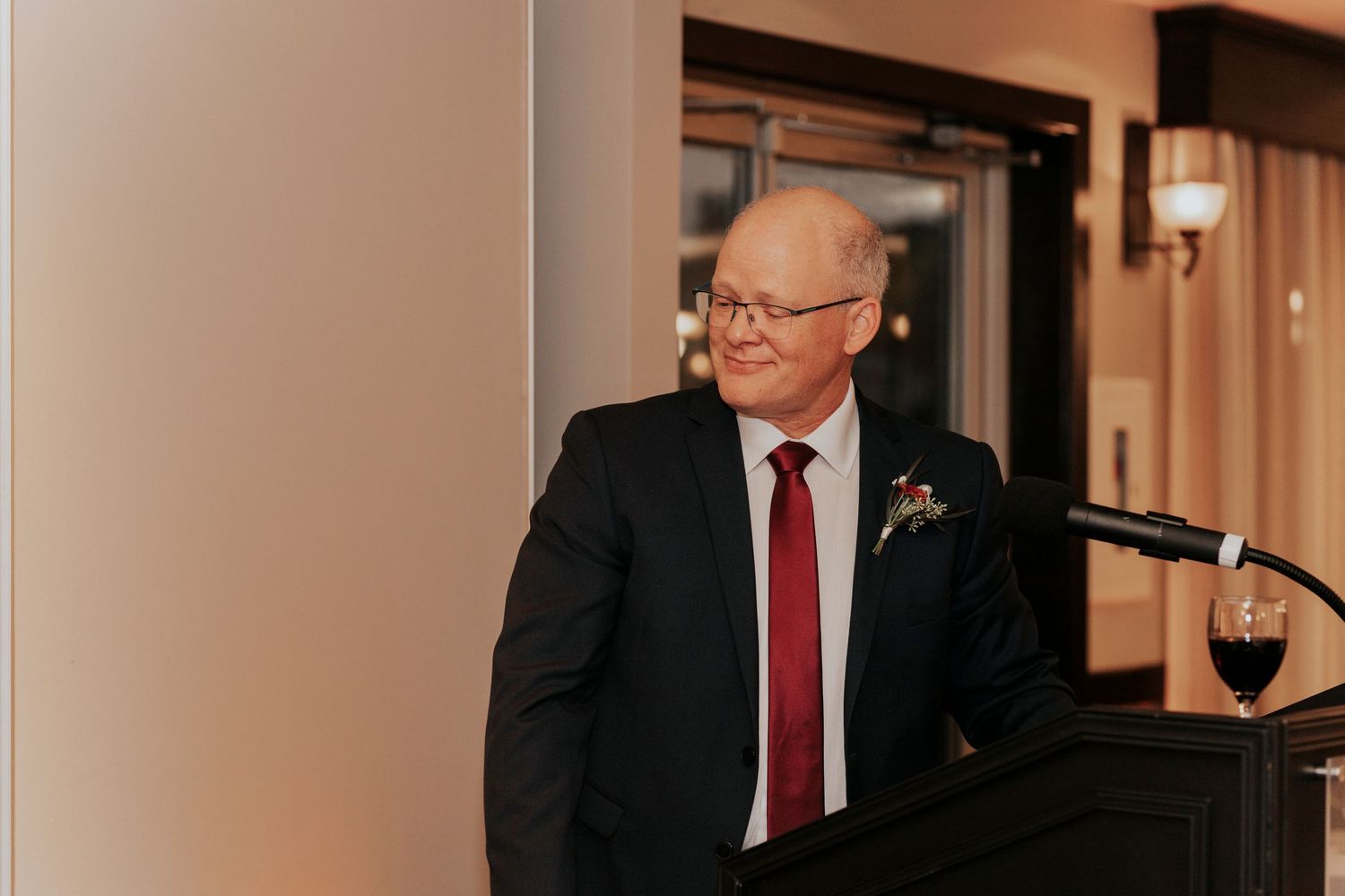 Speaker at wedding reception podium wearing dark suit and red tie.