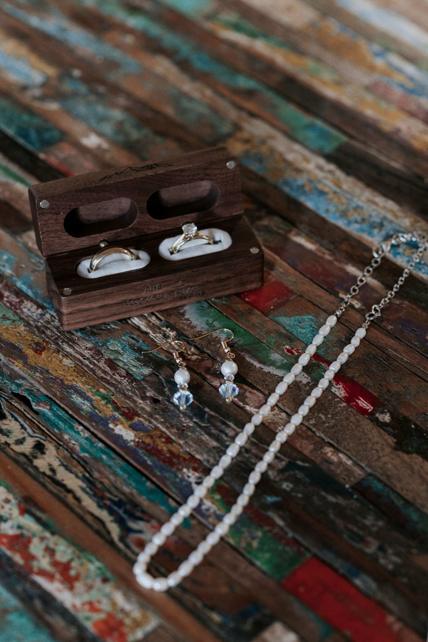 Wedding rings rest in a wooden box alongside delicate pearl jewelry on a distressed multicolored wooden surface.