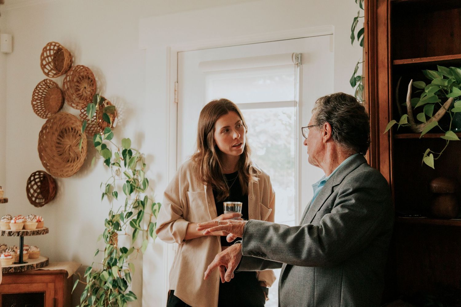 Two people having an animated conversation while standing in a cozy room decorated with woven baskets and trailing plants.