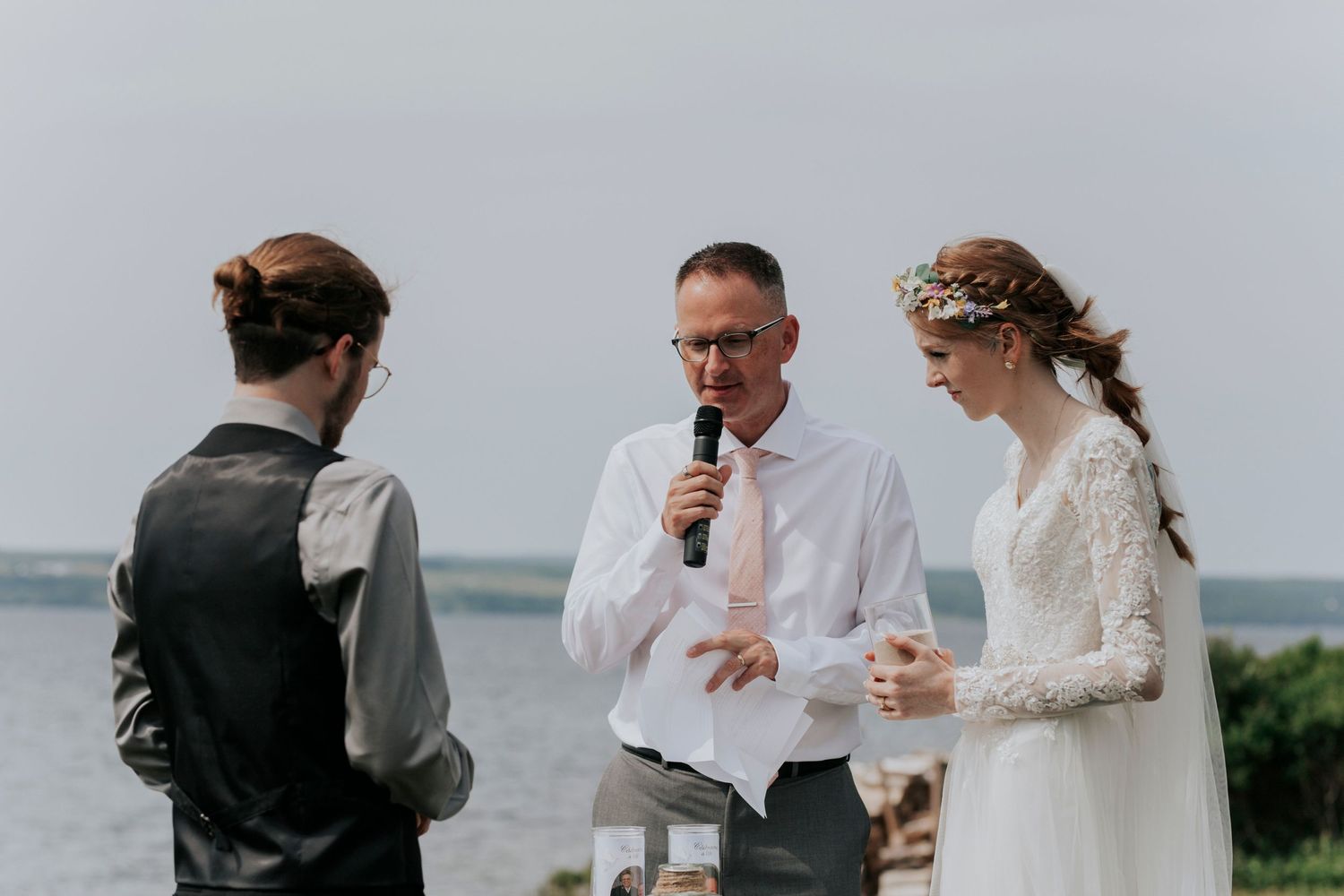 An outdoor wedding ceremony takes place on a misty coastline with an officiant speaking into a microphone.