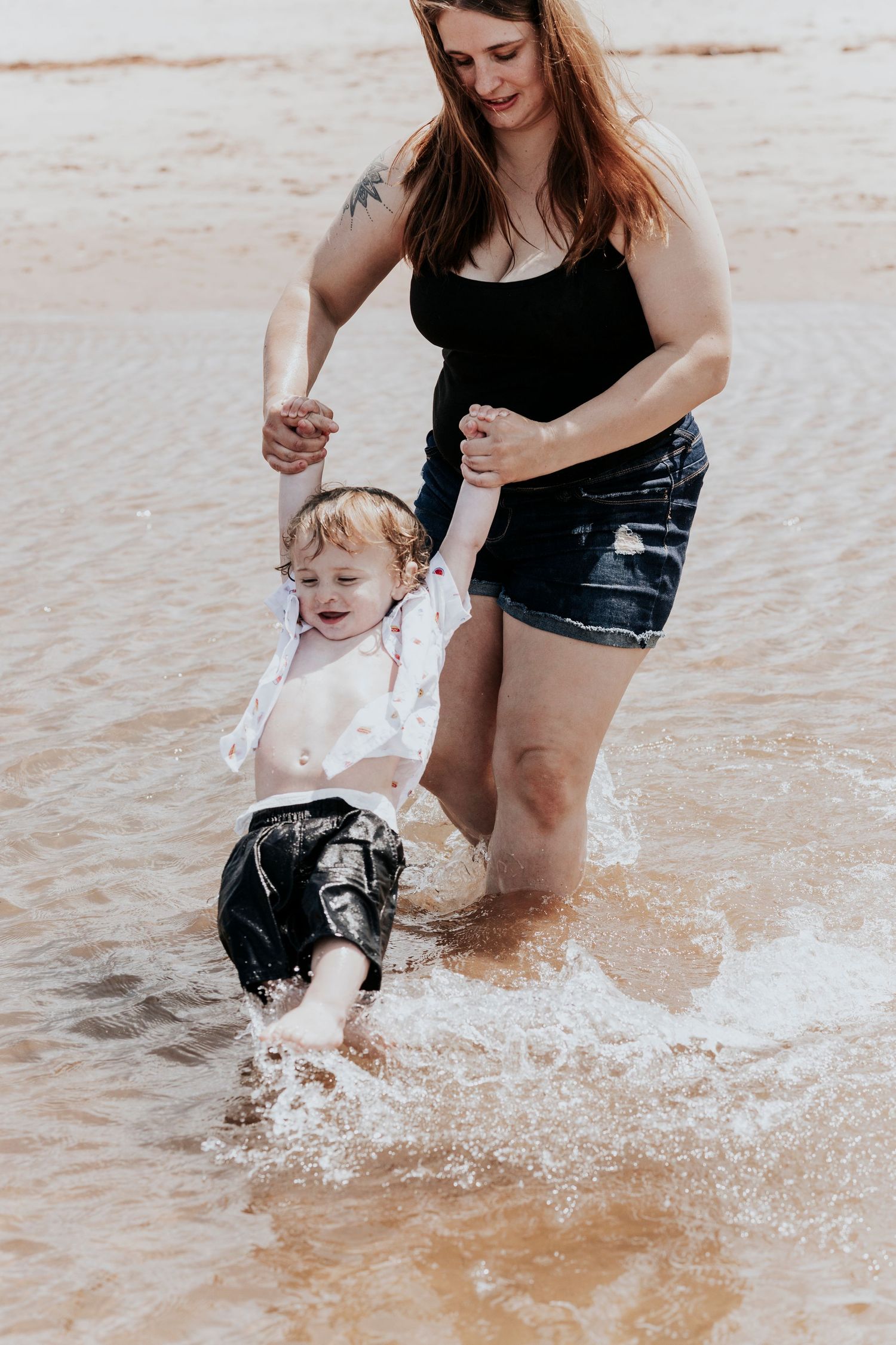Mother holding toddler's hands as he splashes joyfully in shallow ocean waves on a sandy beach.