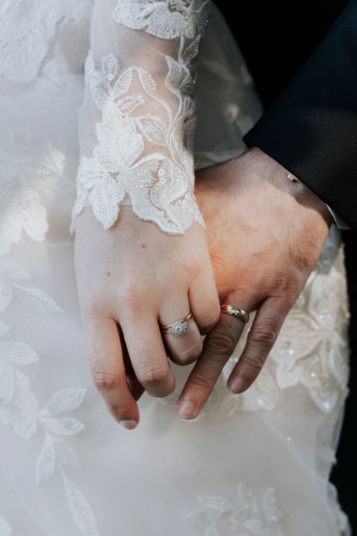 Close-up of newlyweds' hands showing wedding rings with delicate lace detail from the bride's dress.