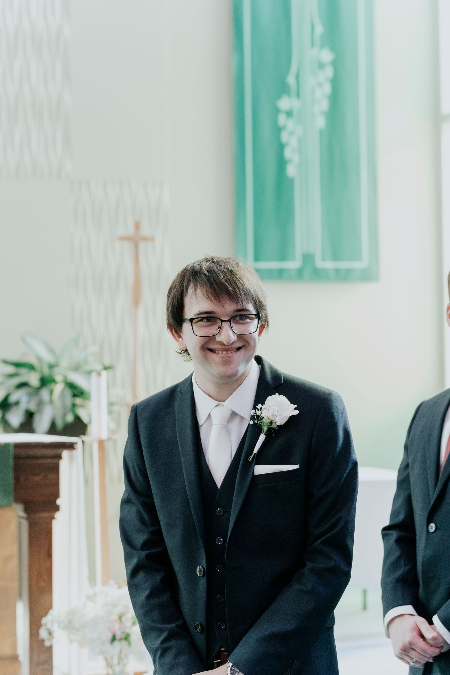 A groom wearing glasses and black suit with white boutonniere smiles at altar in church with mint green decor.