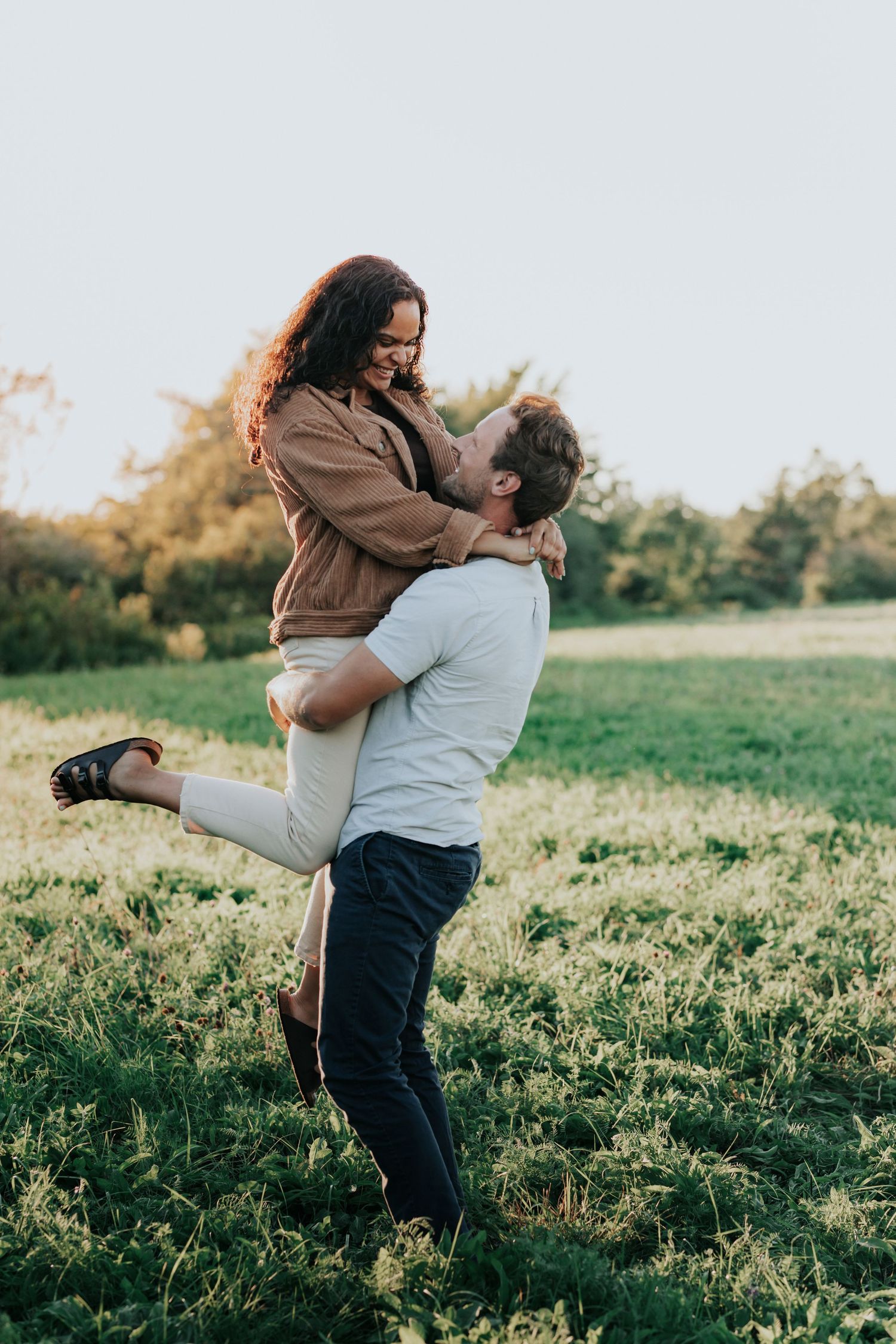 Man lifting woman in a grassy field at sunset during a romantic outdoor engagement session.