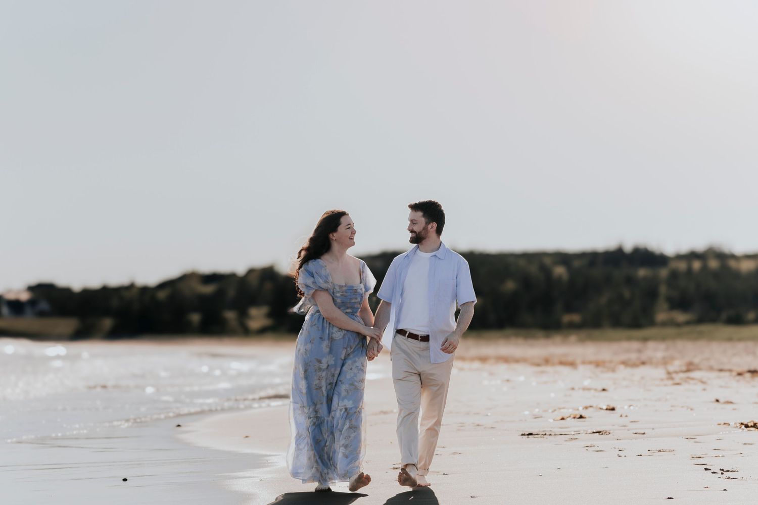 Couple walking hand in hand on sunny beach, smiling at each other in casual summer outfits.