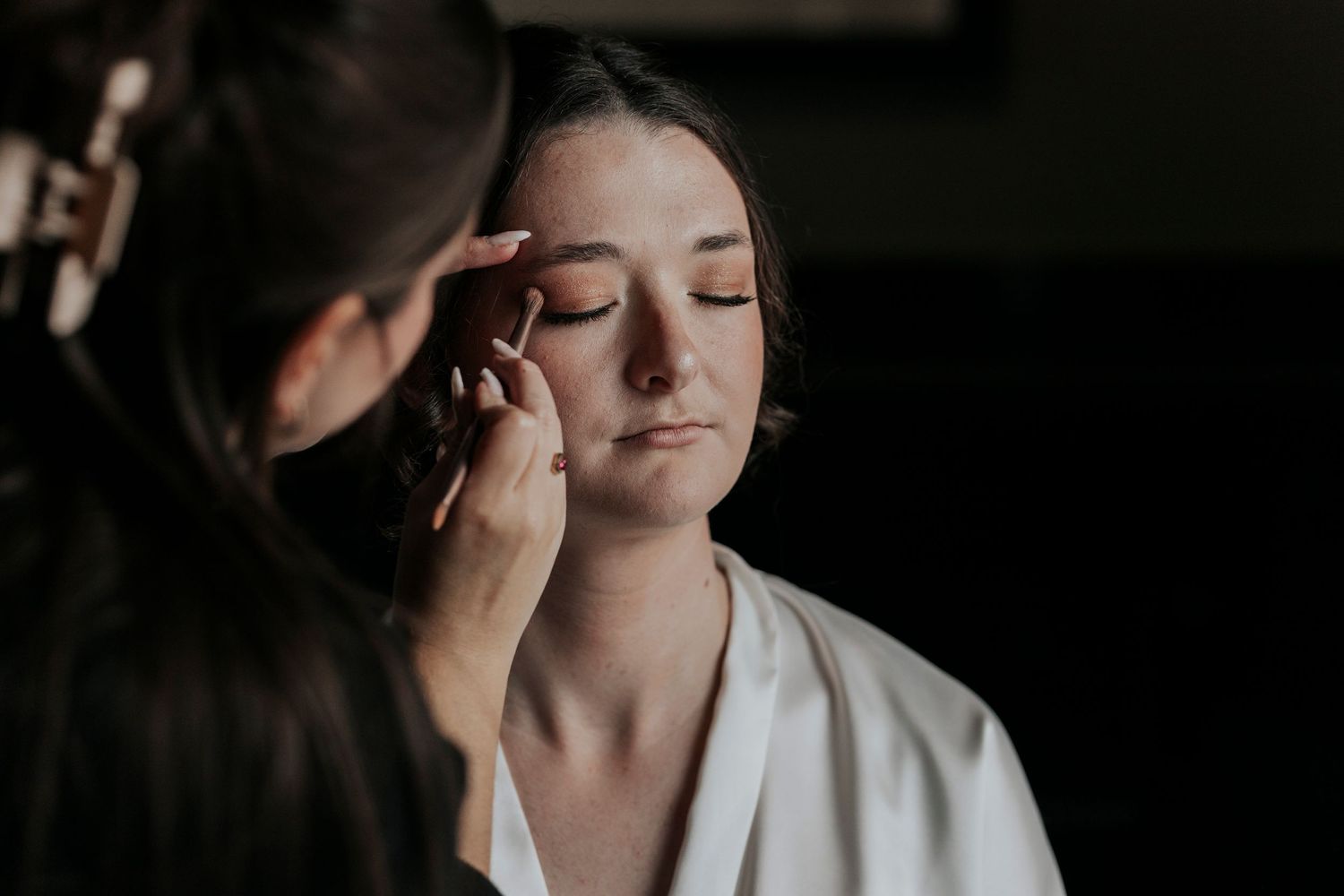 A makeup artist applies wedding day cosmetics in soft, dramatic lighting.