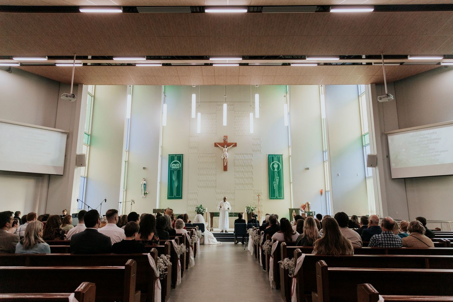 A modern Catholic church interior with congregation seated in pews, facing an illuminated altar with crucifix and green banners.