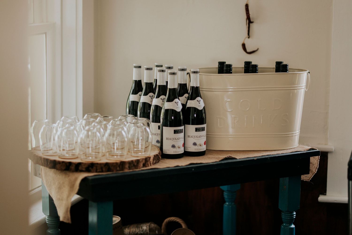 A rustic bar setup with glass bottles, stemware, and a white ice bucket on a dark wooden surface.
