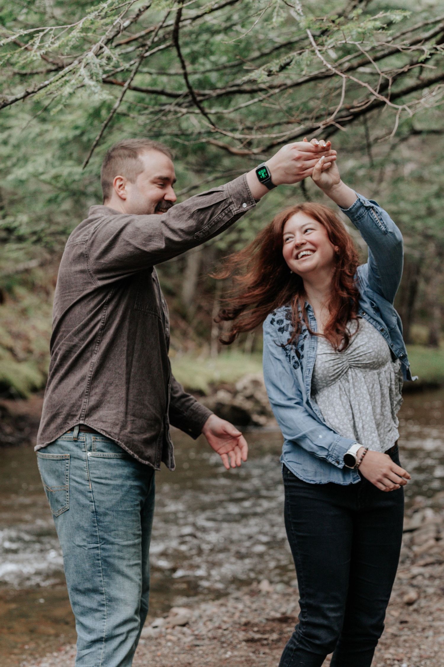 Couple dancing playfully on rocky riverbank in forest, man spinning laughing redhead woman outdoors.