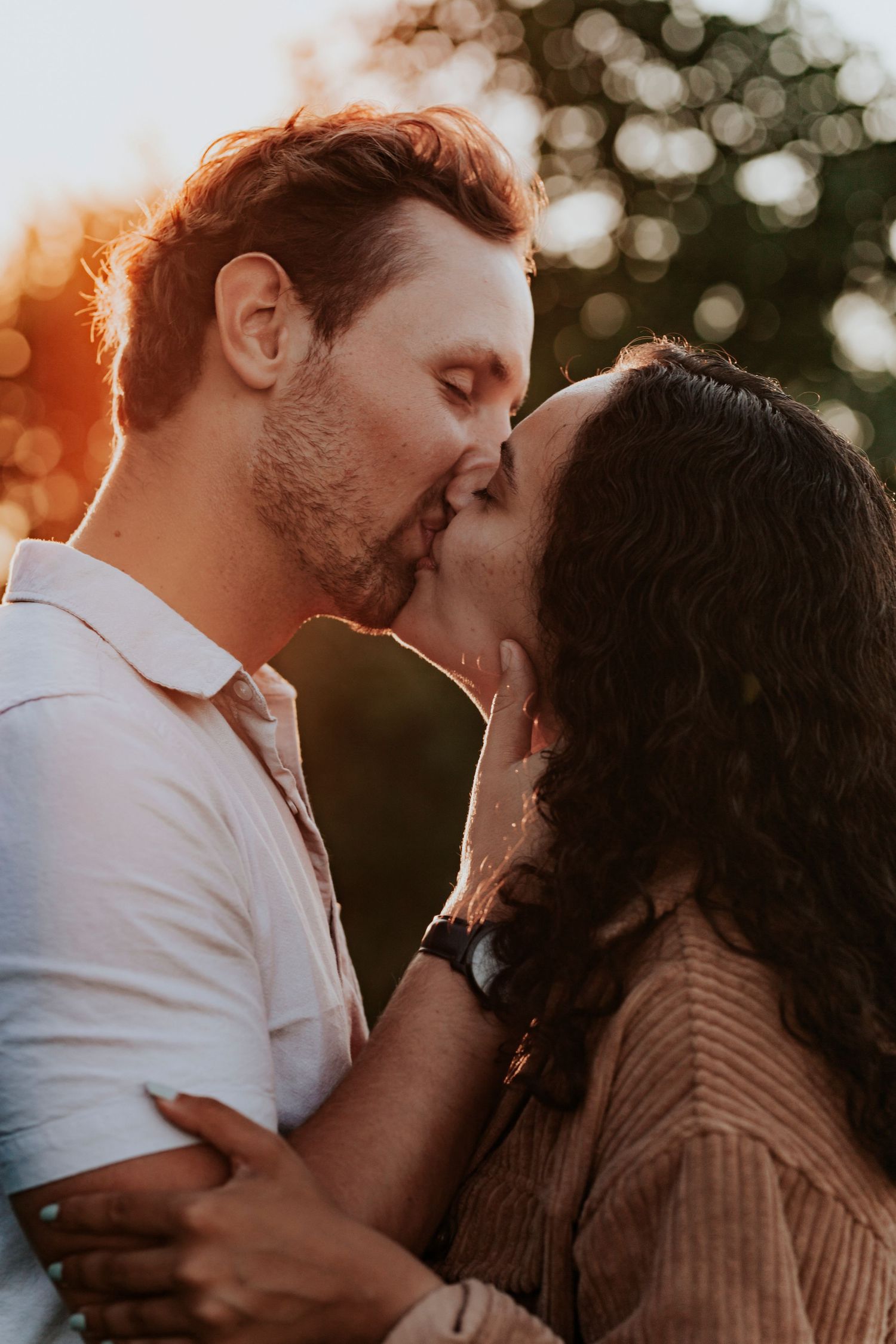 Couple sharing a romantic kiss outdoors with warm golden bokeh sunset light in the background.