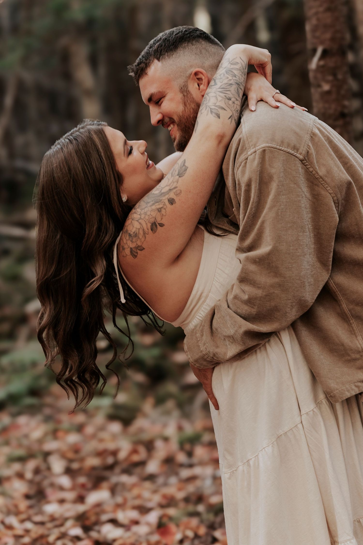 Couple embraces and laughs together in autumn forest, woman with tattoos in white dress, man in tan jacket.