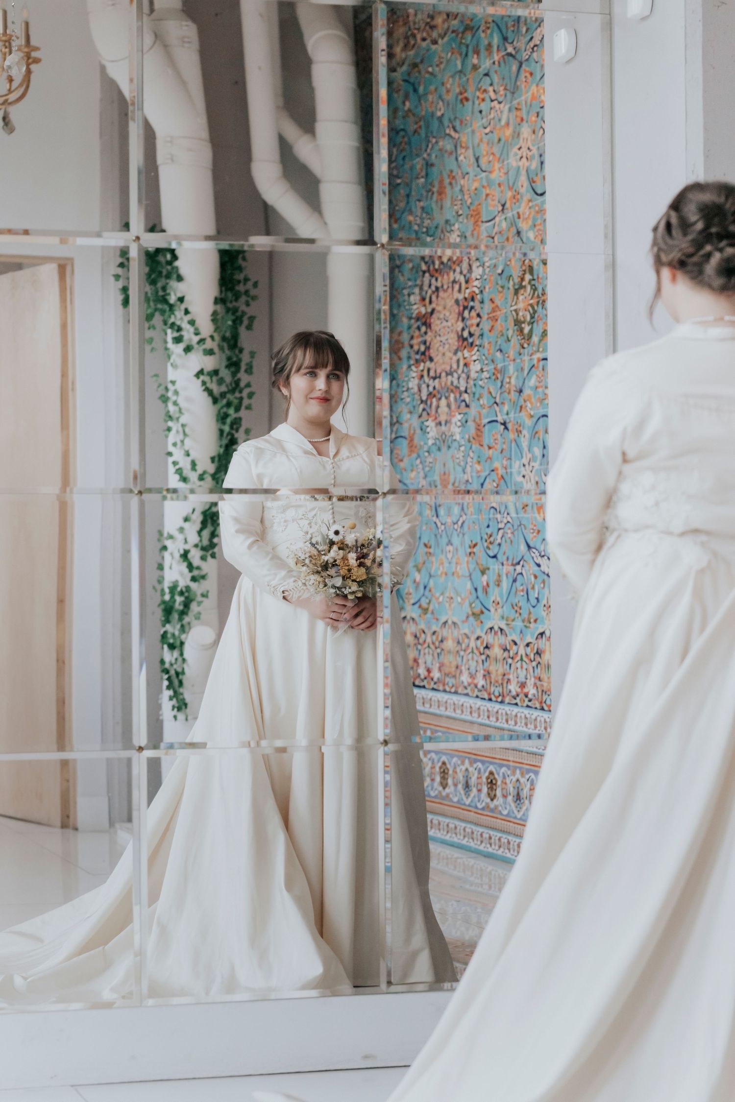 Bride in a long-sleeve ivory gown holds a bouquet while admiring her reflection in a decorative mirror.