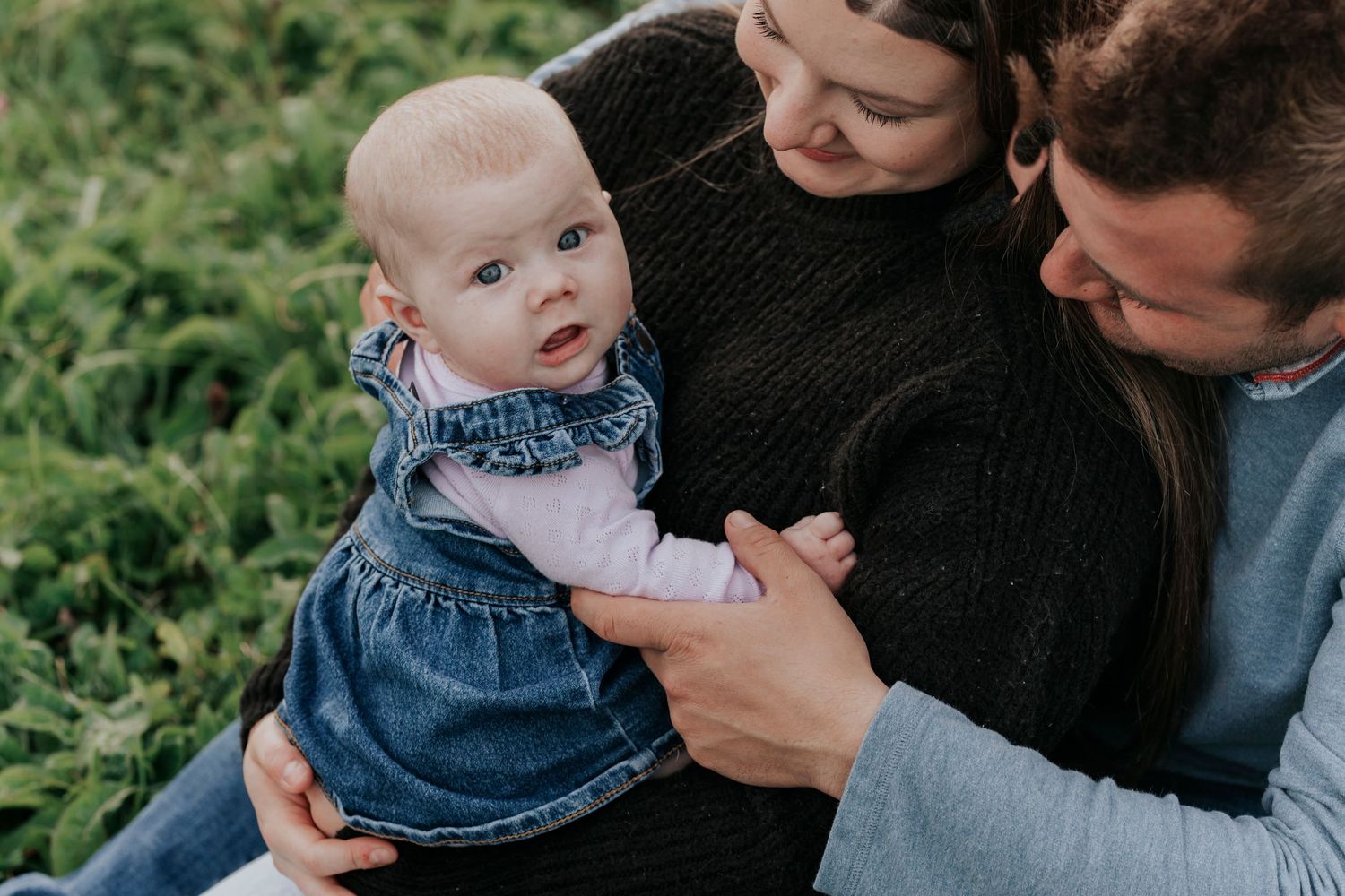 Parents lovingly holding baby girl in denim dress outdoors.