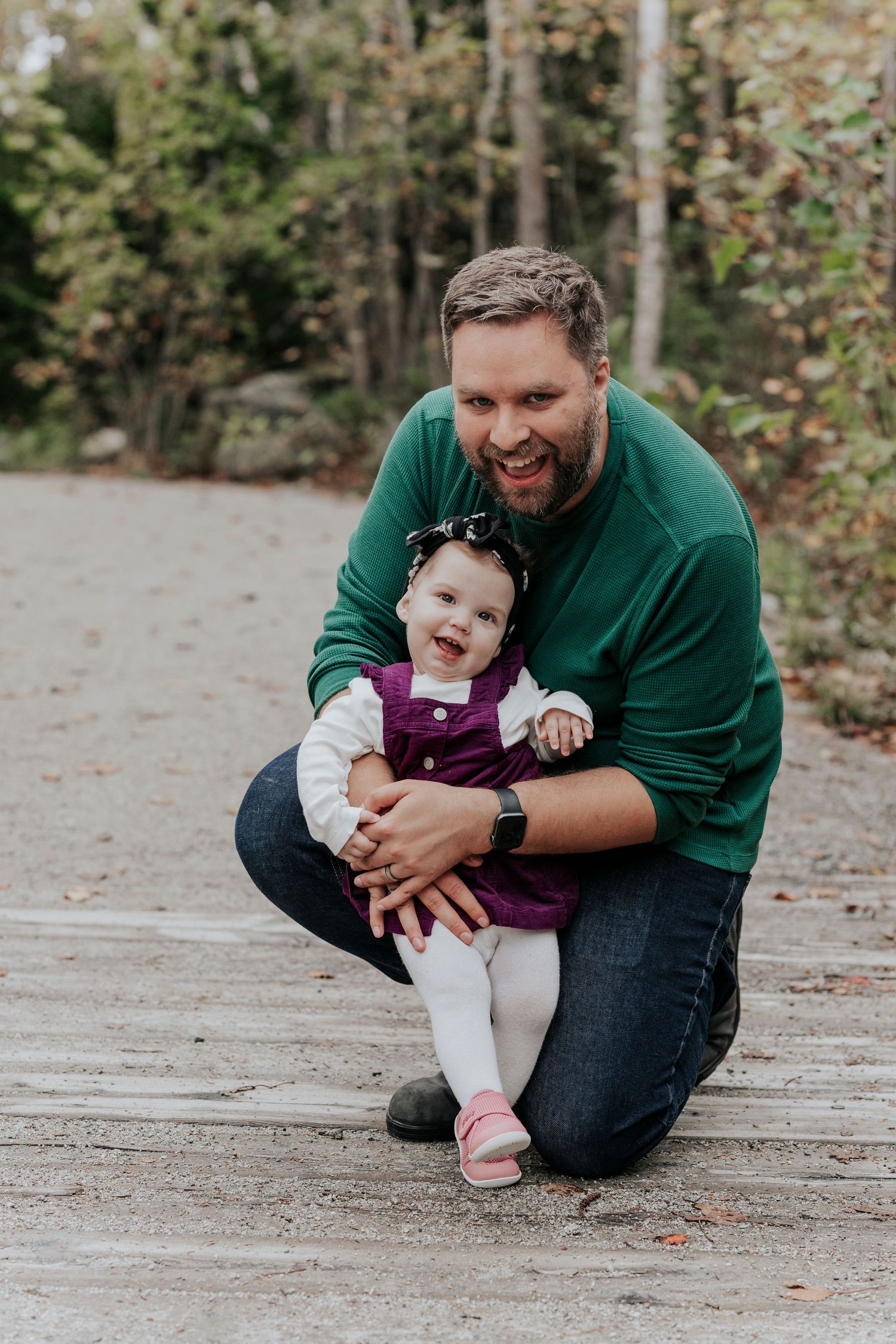 Bearded father in green sweater crouching and hugging baby girl on woodland path.