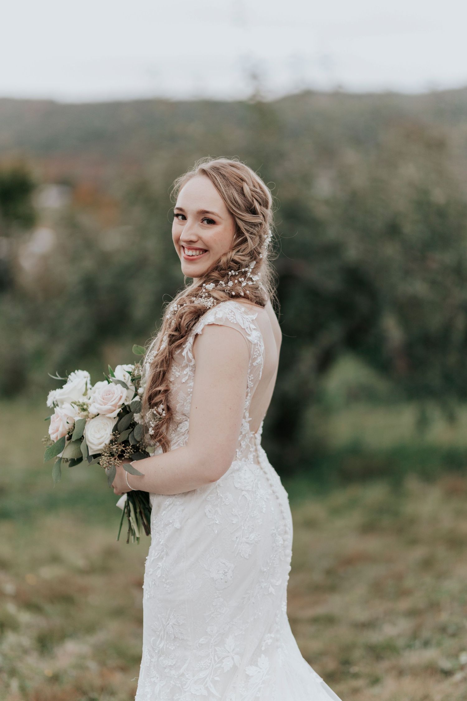 A smiling bride in a lace mermaid gown holding a white bouquet in an outdoor orchard setting.