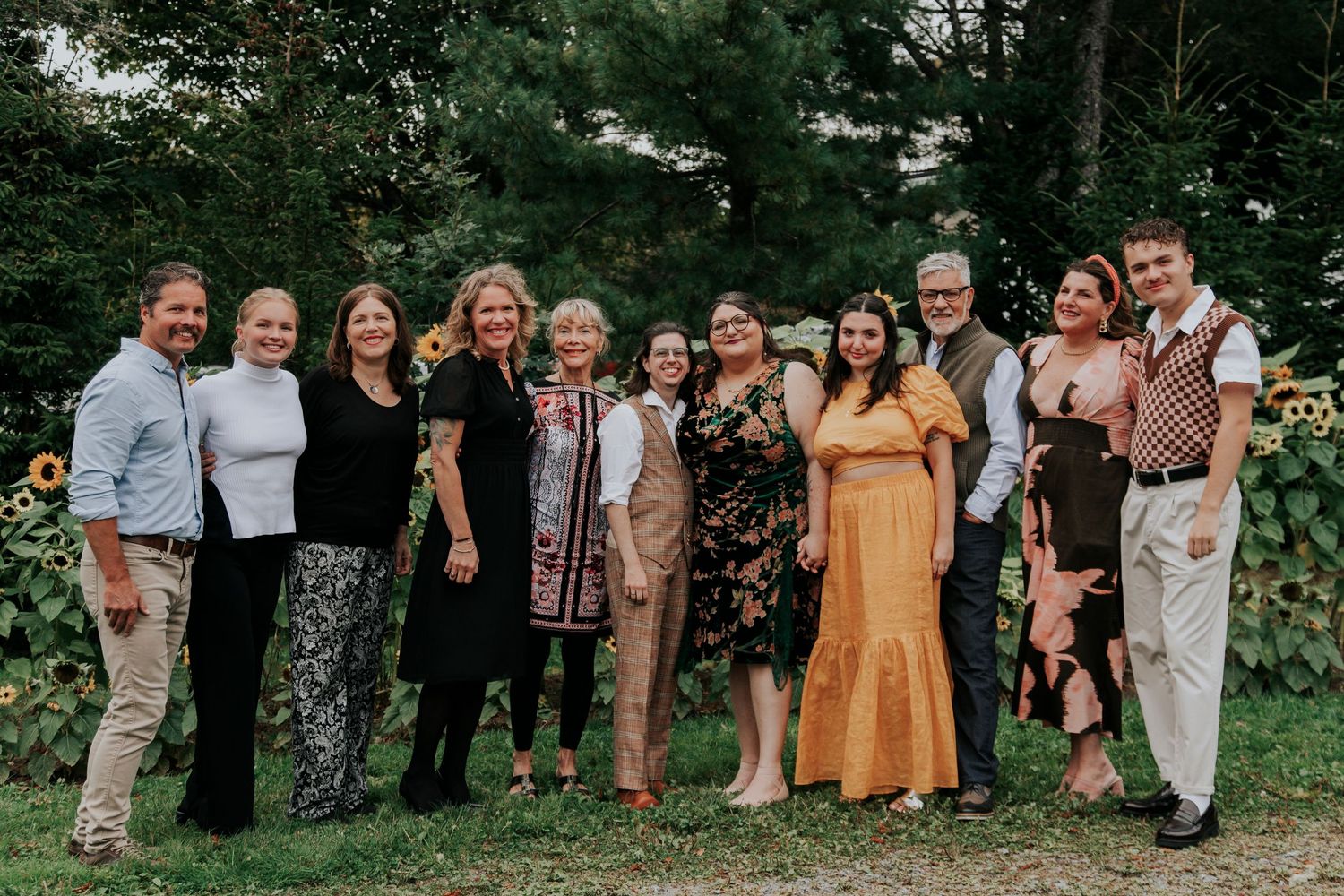 A group of eleven wedding guests smiling together outdoors near a sunflower garden.