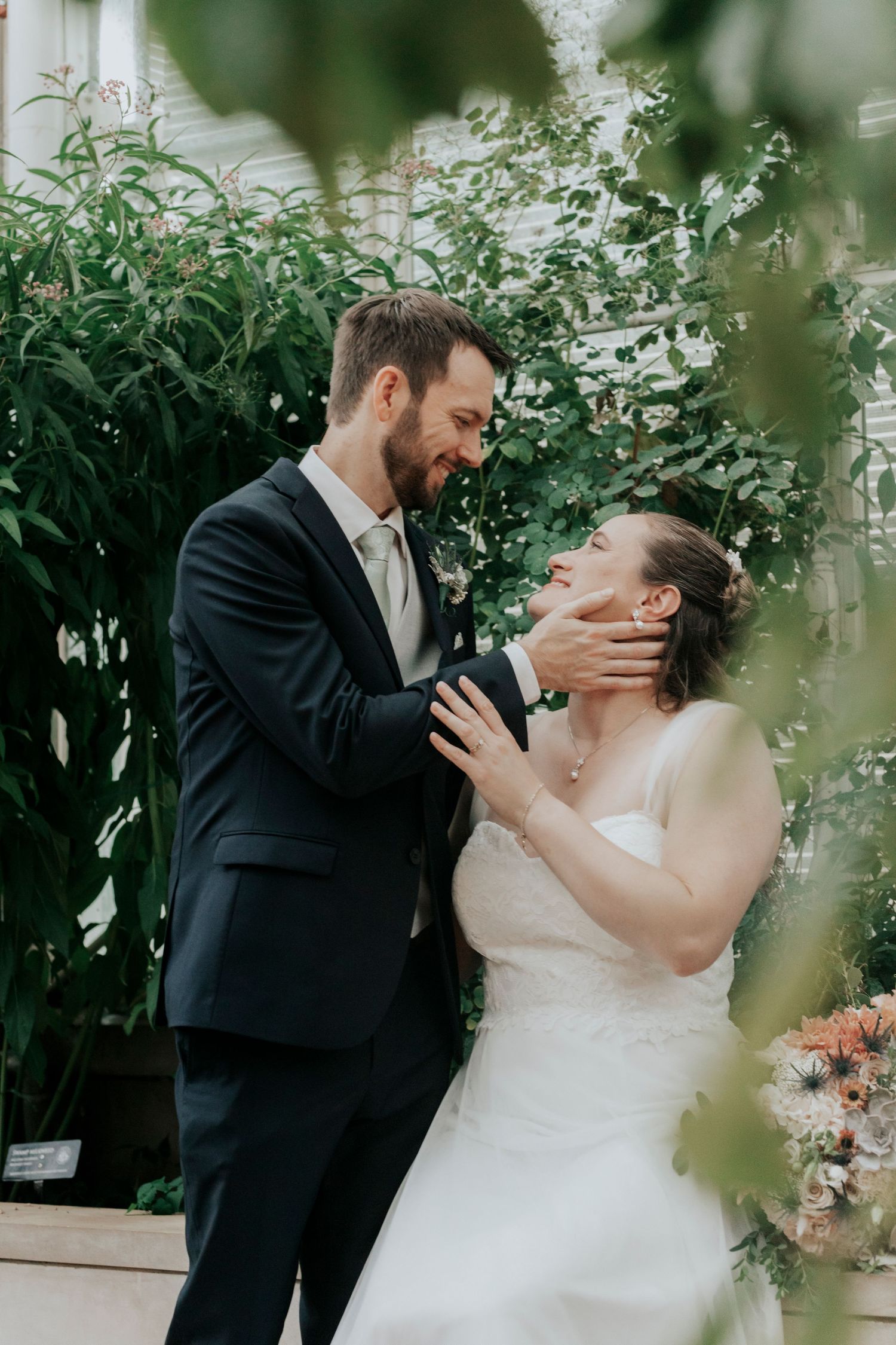 Groom gently cups brides face as they share a joyful moment surrounded by lush greenhouse greenery.