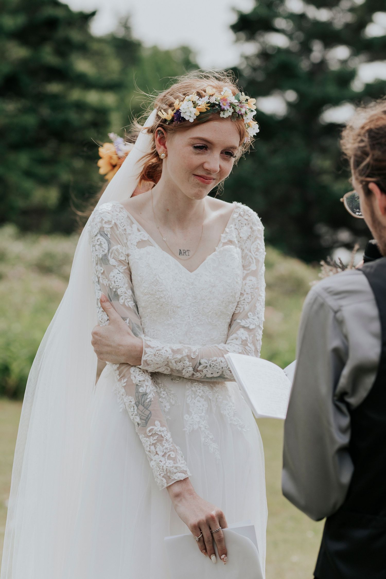 A bride wearing a delicate lace long-sleeved wedding gown and floral crown stands outdoors during her wedding ceremony.