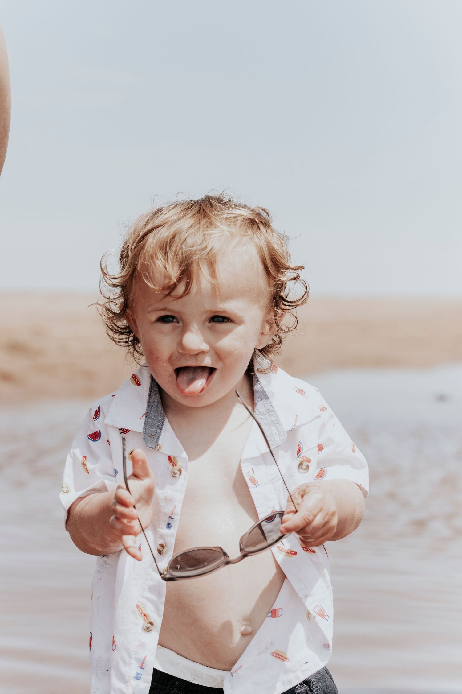 Cheerful curly-haired toddler in open patterned shirt holding sunglasses on a sandy beach.