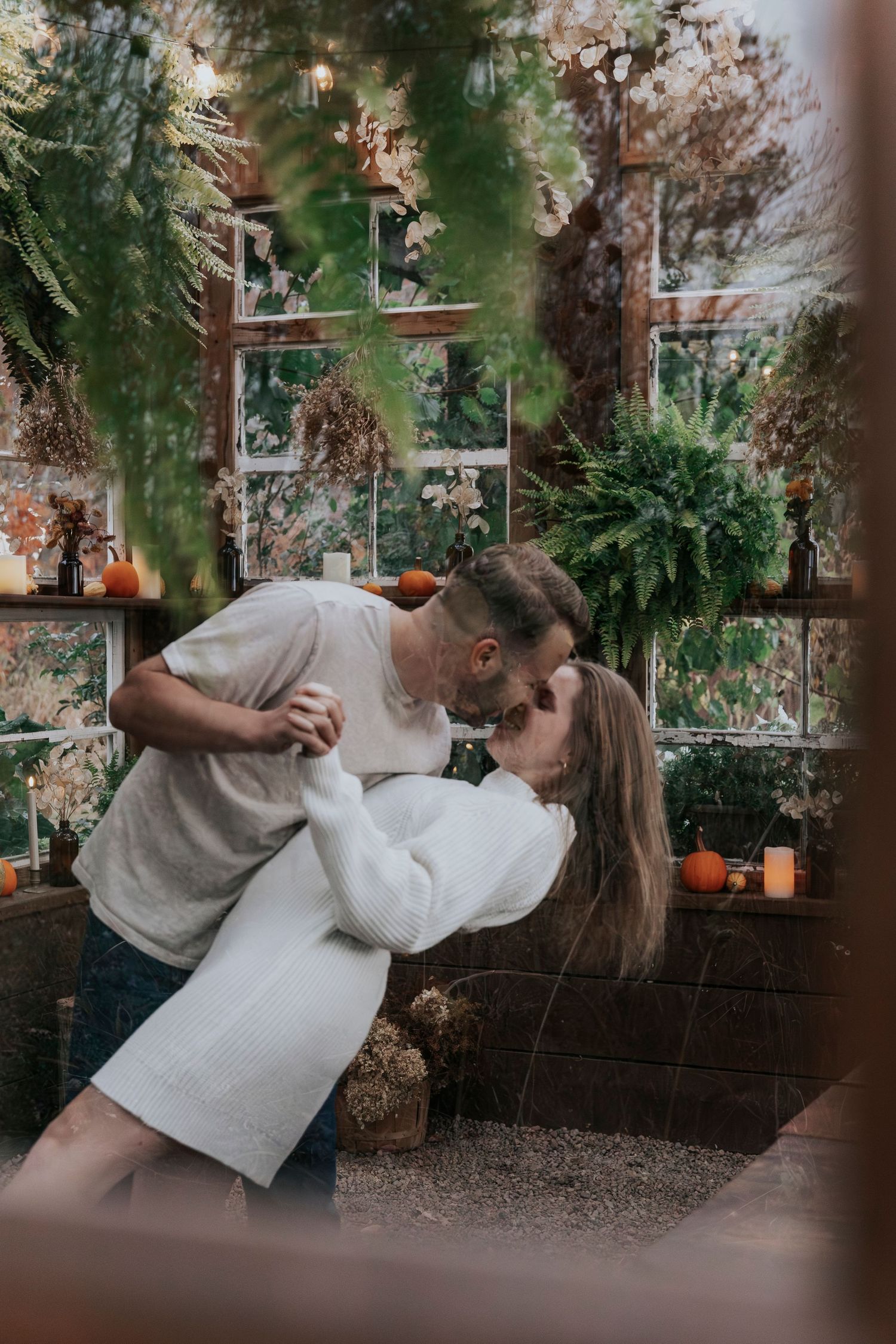 Couple sharing a romantic dip kiss inside a lush greenhouse with candles and plants.