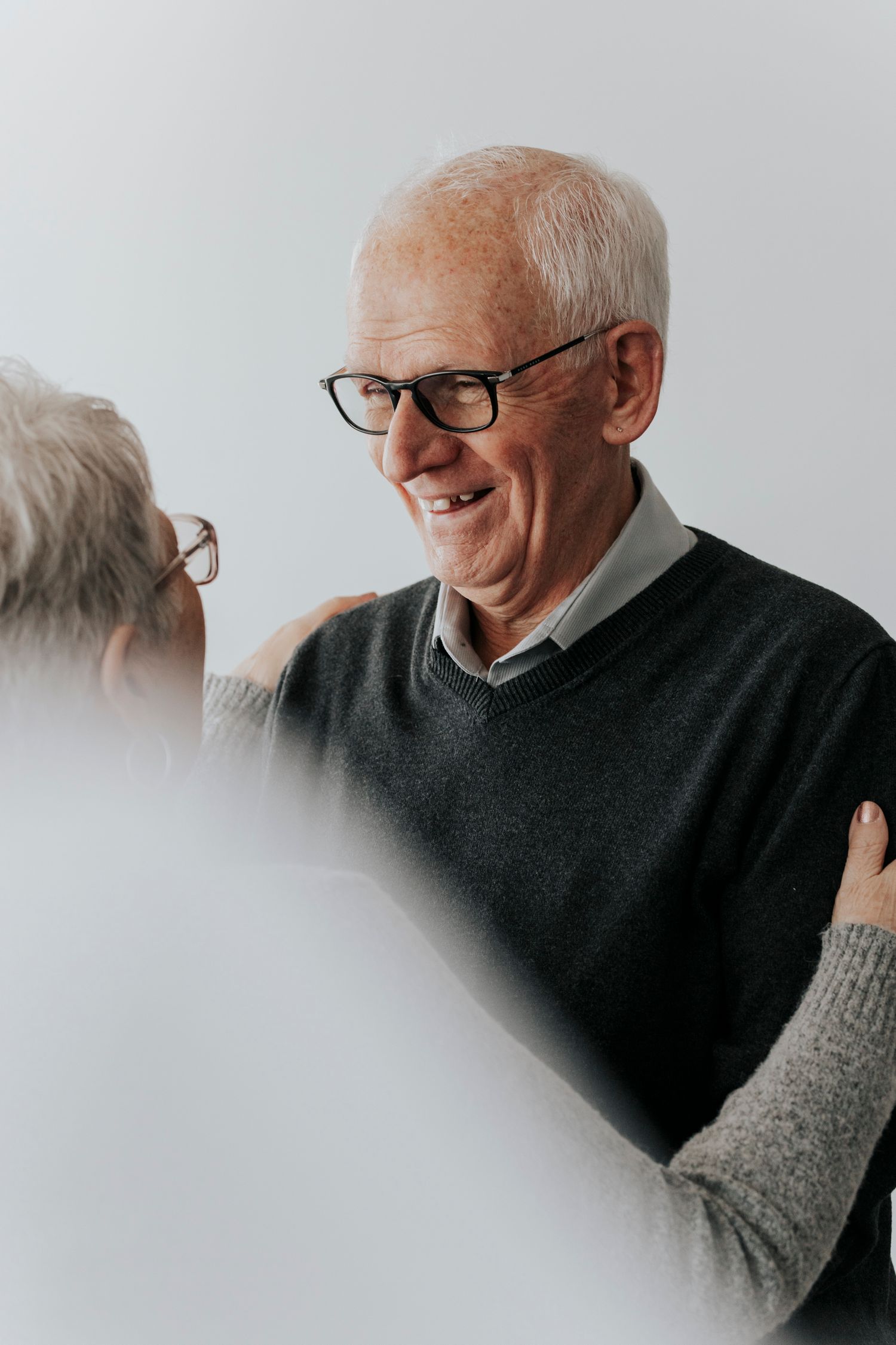 Smiling elderly man with glasses wearing a dark sweater being embraced in a bright indoor setting.