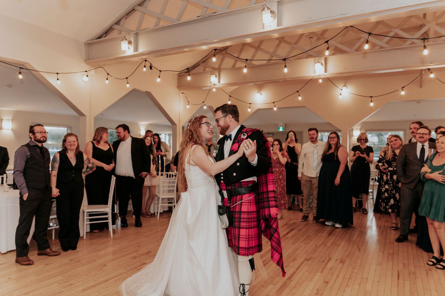 Bride and groom sharing first dance in rustic venue with string lights and wedding guests watching.