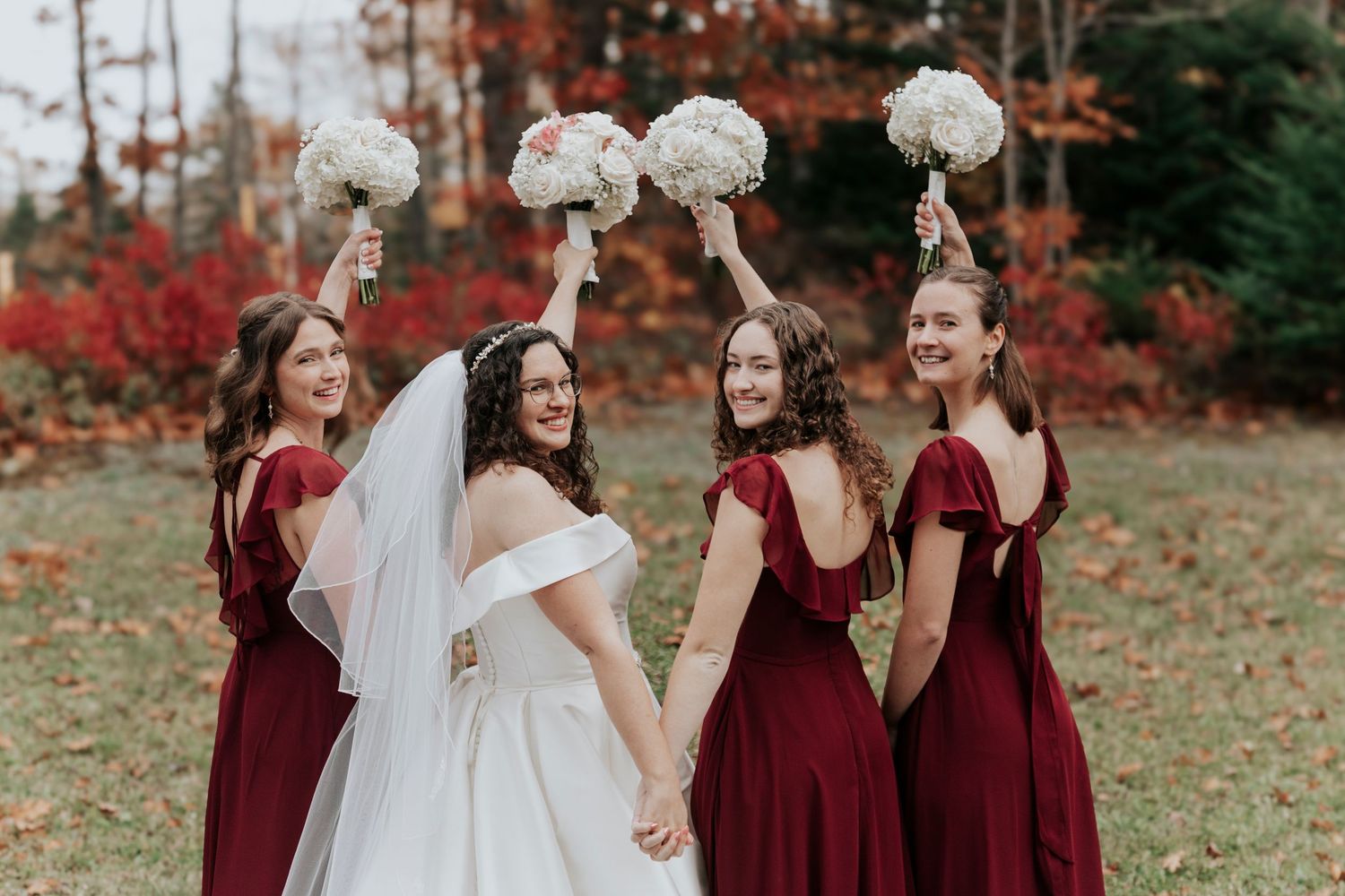 Bride in white gown with bridesmaids in burgundy dresses raising bouquets in fall setting.