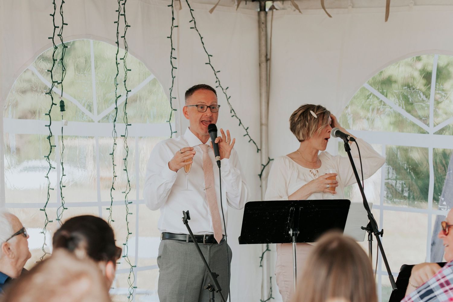 Two people give speeches into microphones at a wedding reception under an outdoor tent with hanging greenery decor.