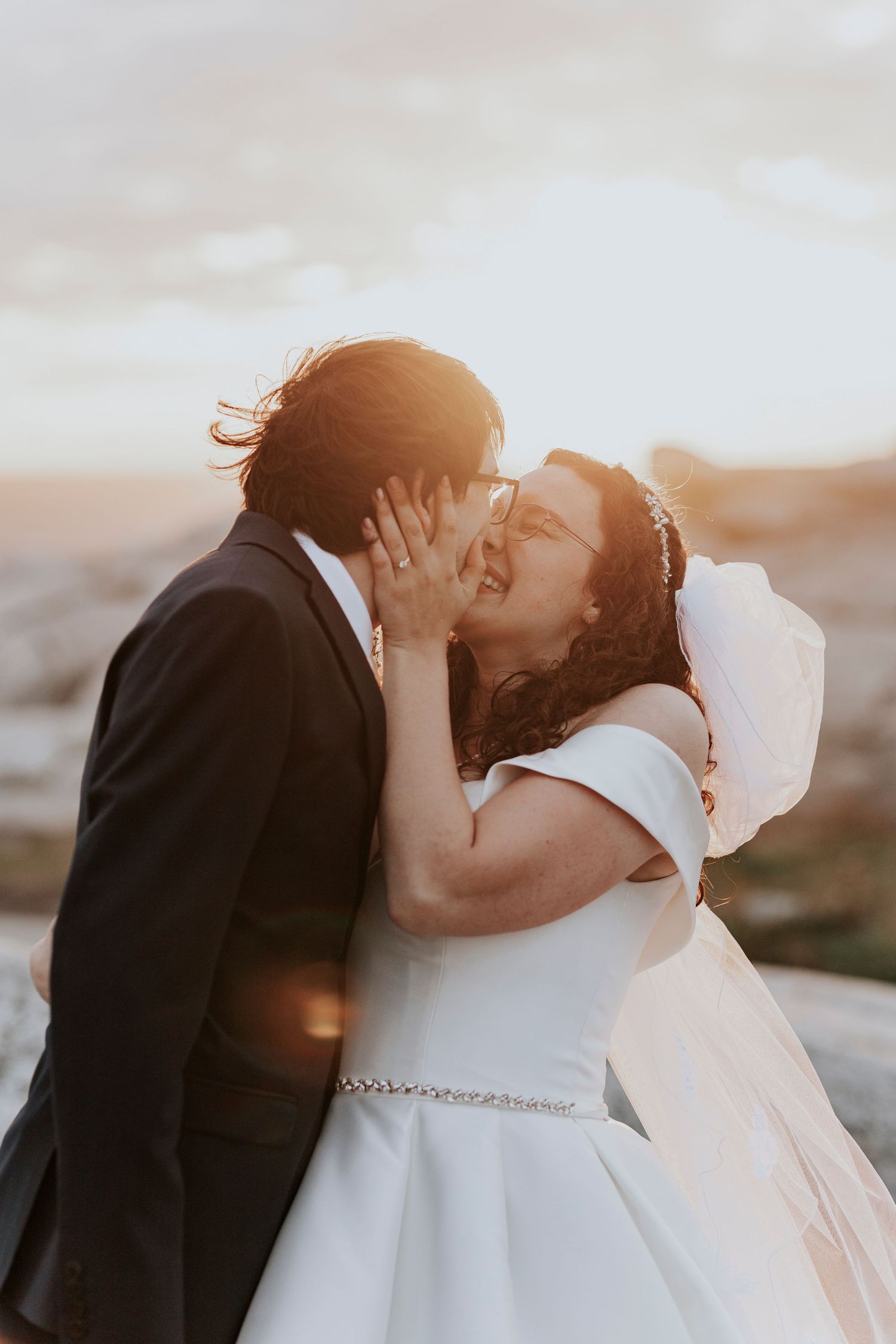 Romantic sunset wedding photo with couple sharing intimate kiss outdoors at scenic overlook.