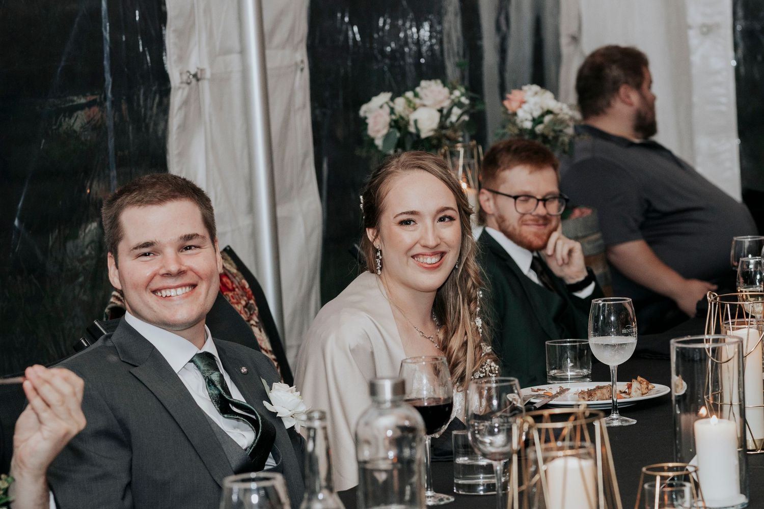 Newlyweds and wedding party smile during reception dinner under white tent decor and string lights.