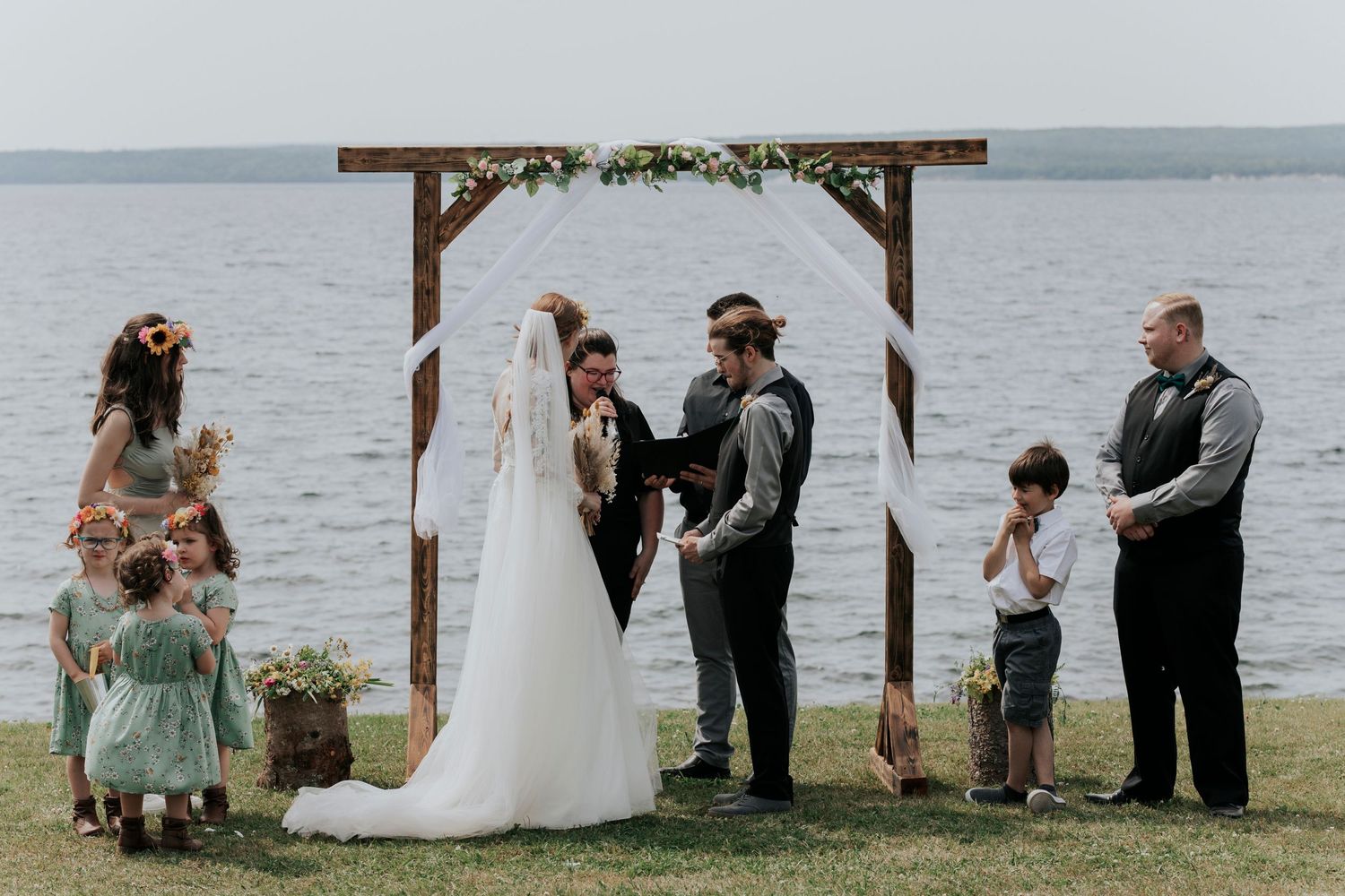 A lakeside outdoor wedding ceremony with a wooden arch decorated with greenery against a waterfront backdrop.