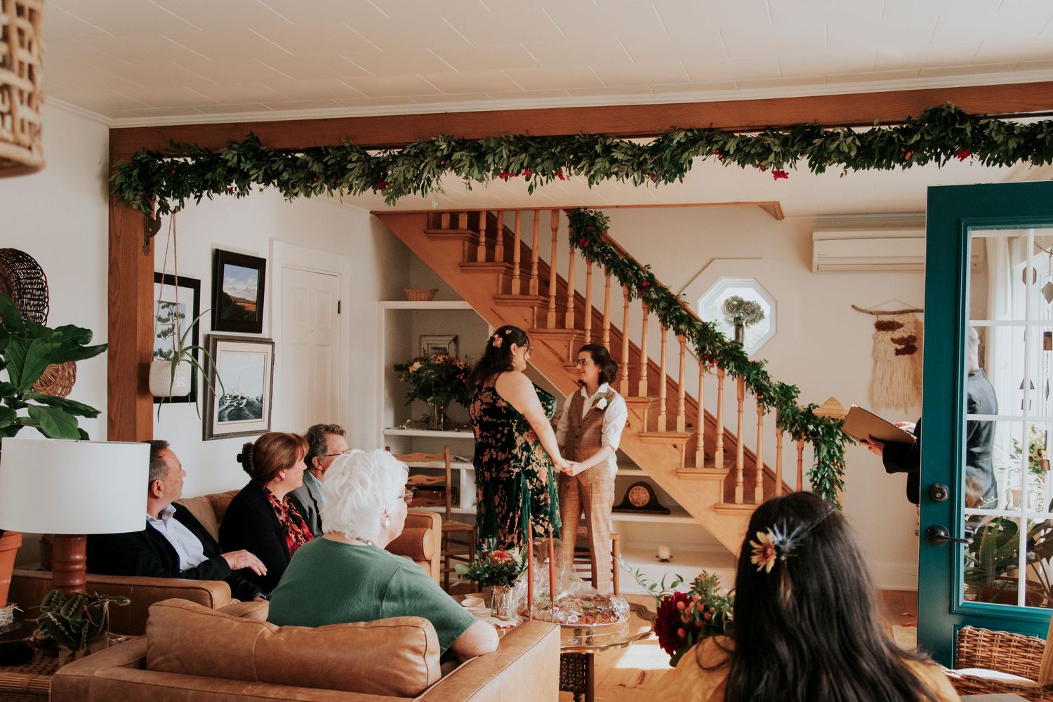 A cozy living room decorated for the an at home wedding with festive greenery on the stair banister and people gathered together.