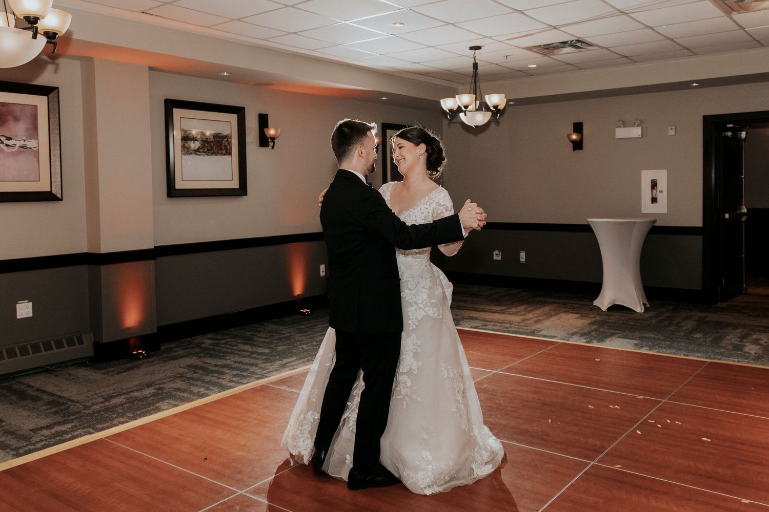 Couple shares their first dance on a wooden floor in a dimly lit reception hall.